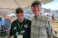 Two smiling men under a tent at an outdoor event, one in green, one in plaid.ght