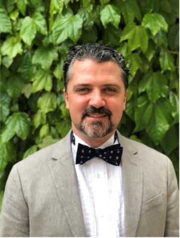 Smiling man with beard in gray suit and patterned bow tie against a green leafy wall.